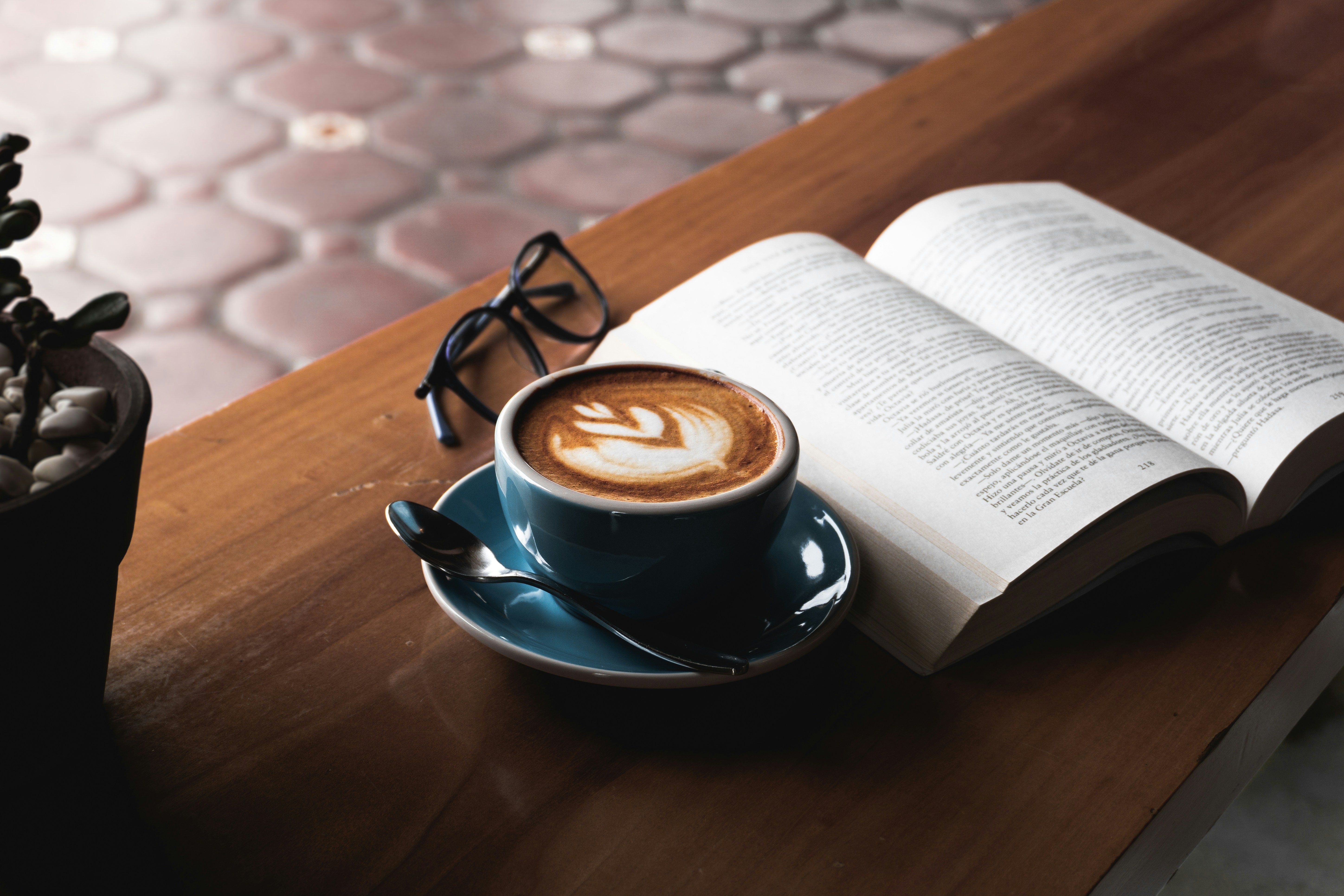 An open book next to a pair of glasses and a latte in a rounded blue cup.