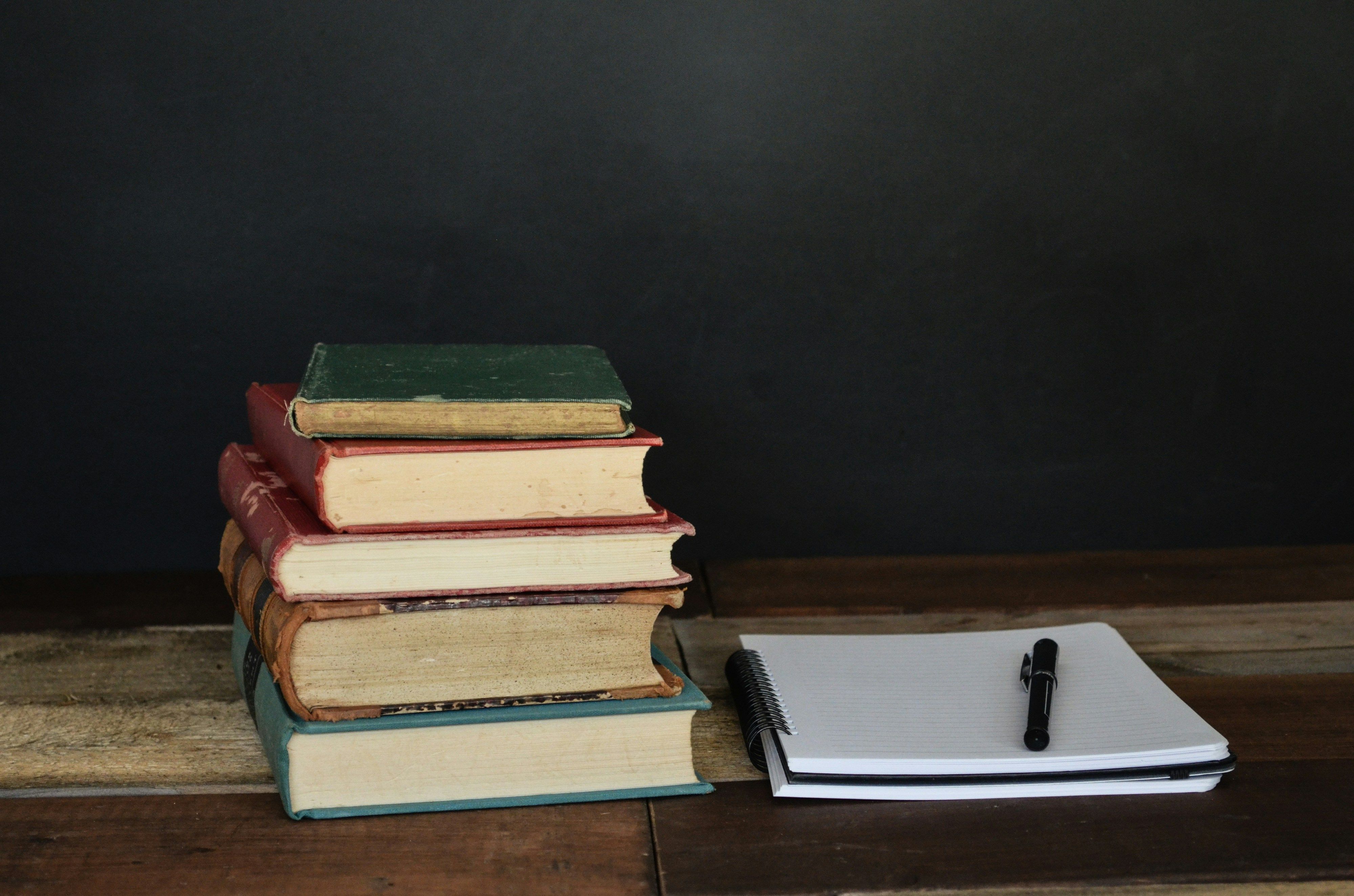 Five books stacked next to a spiral-bound notebook with a pen on top of it.