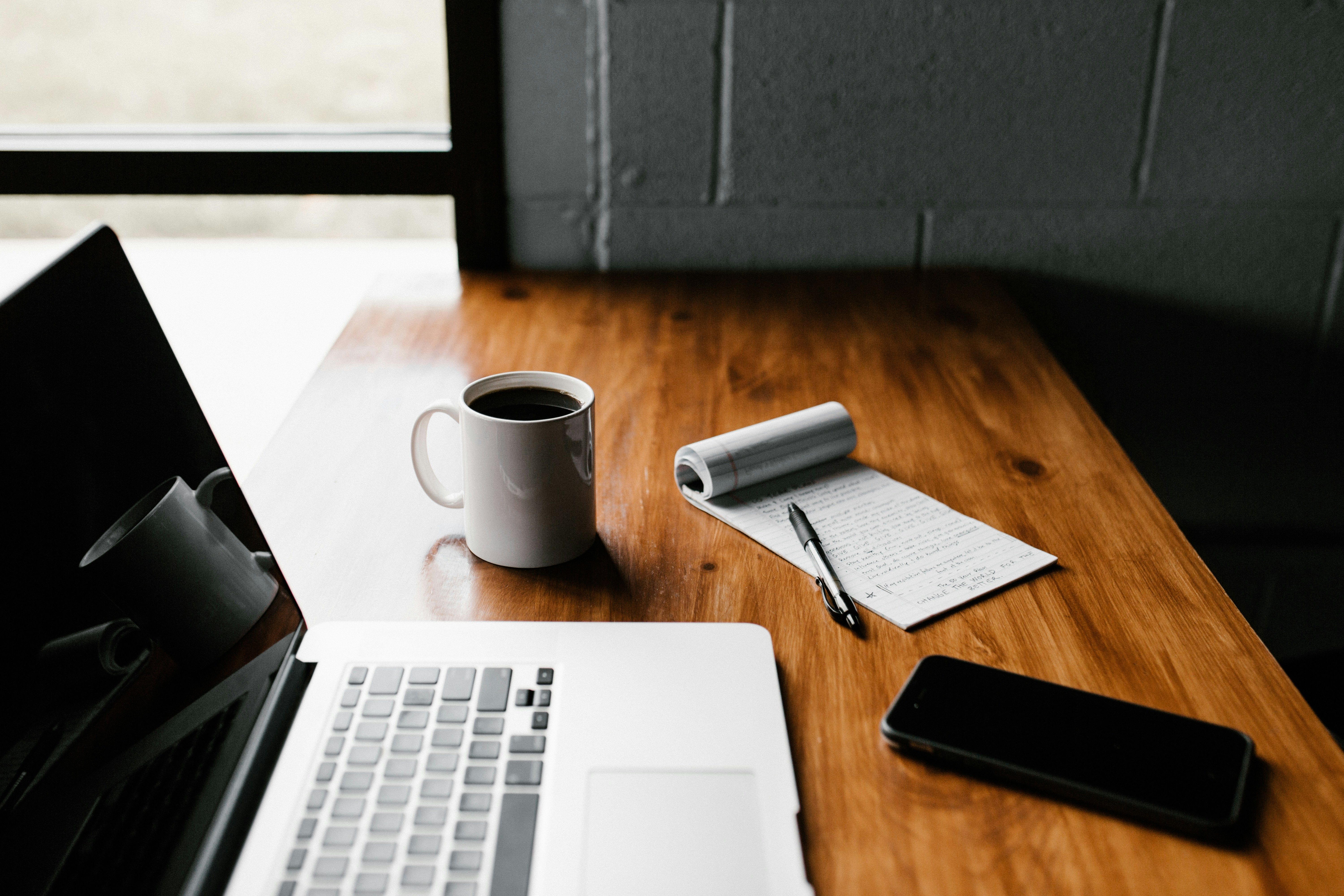 A laptop next to a cup of coffee, a phone, and an open notebook with a pen on top of it.